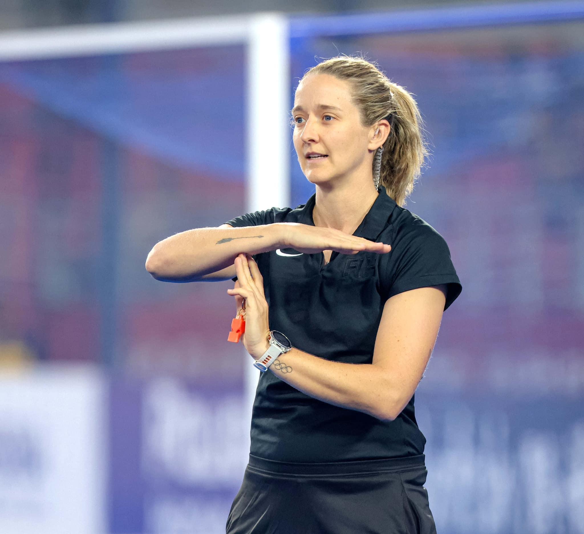 Aleisha Neumann, wearing a black t-shirt and skirt, is standing on a hockey pitch with her arms in a T position, and a whistle in her hand.