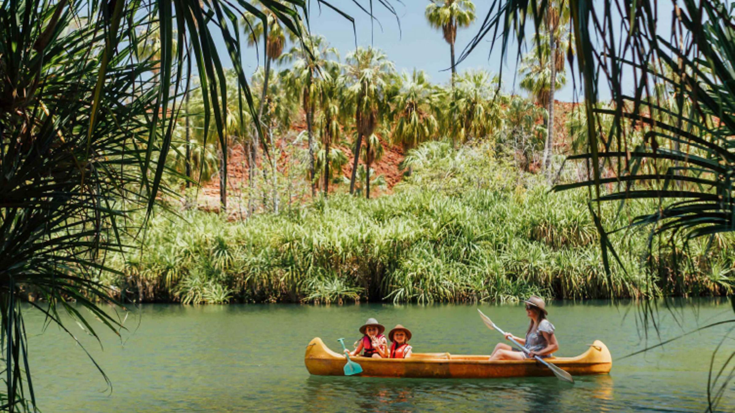 A mother and her two children canoe up a small river with tropical greenery on both shores