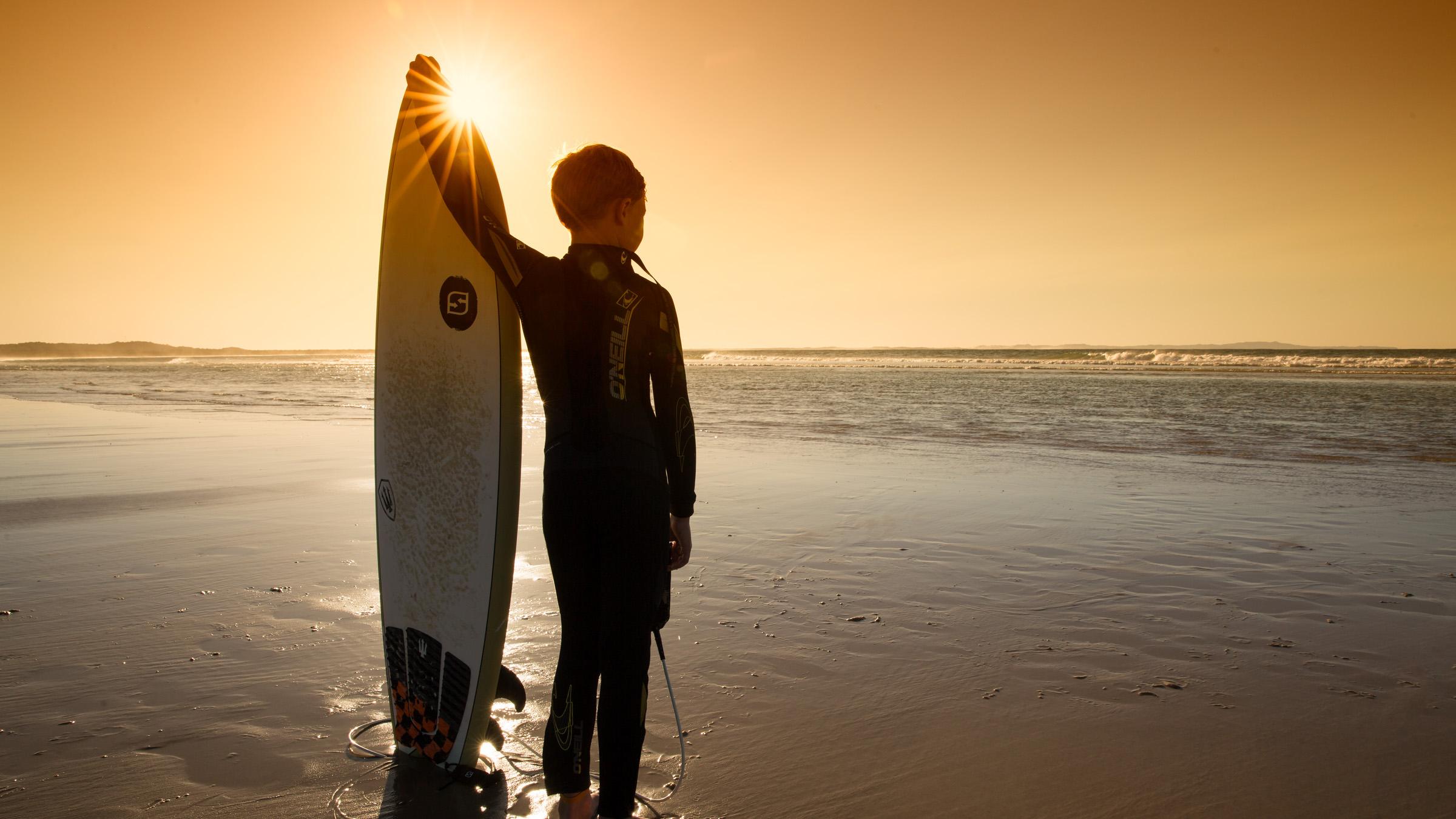 Child surfer on the shore holding a surfboard and looking out to the water