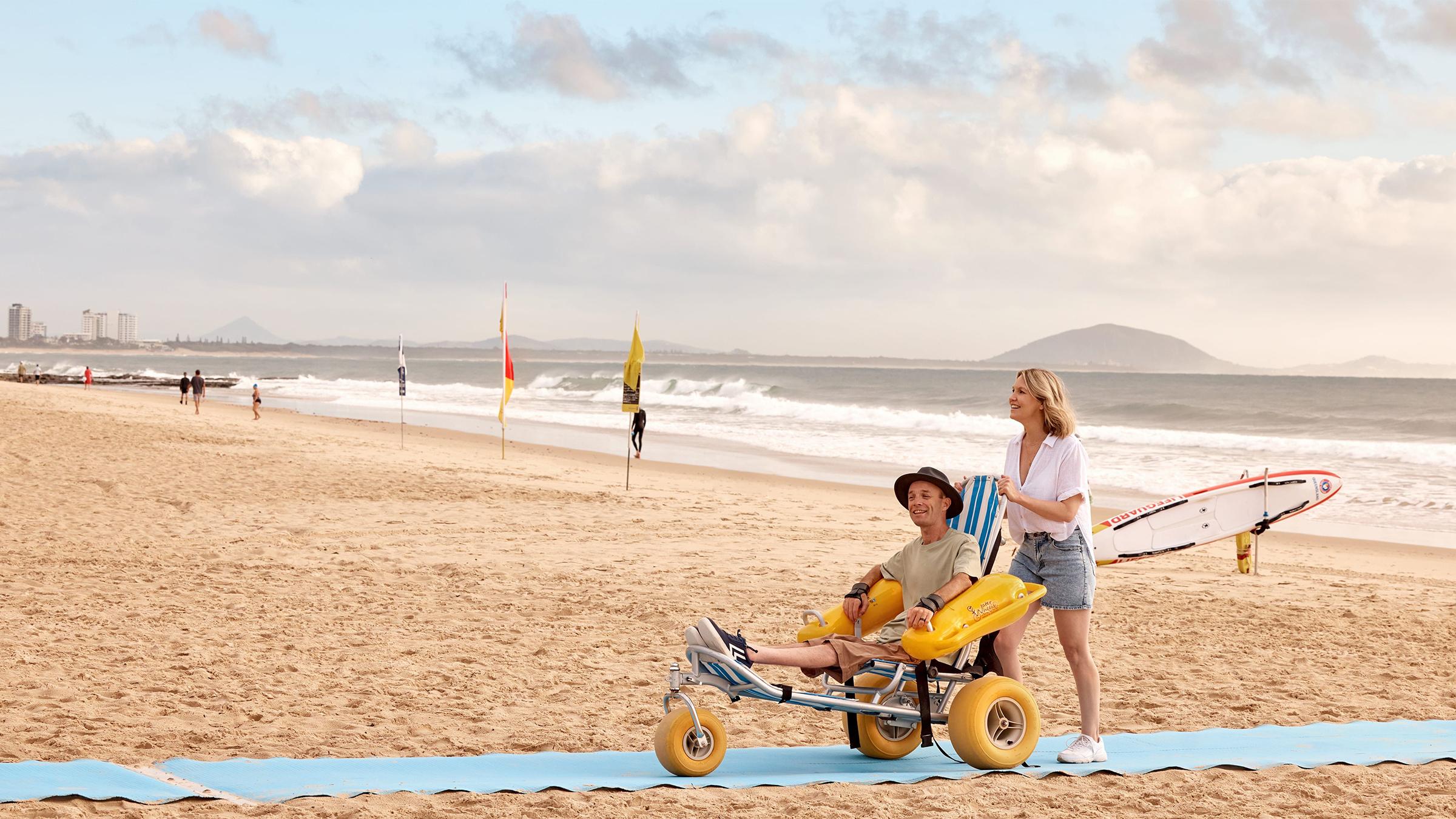 A woman is pushing a man in a wheelchair along a carpet path on the sand.