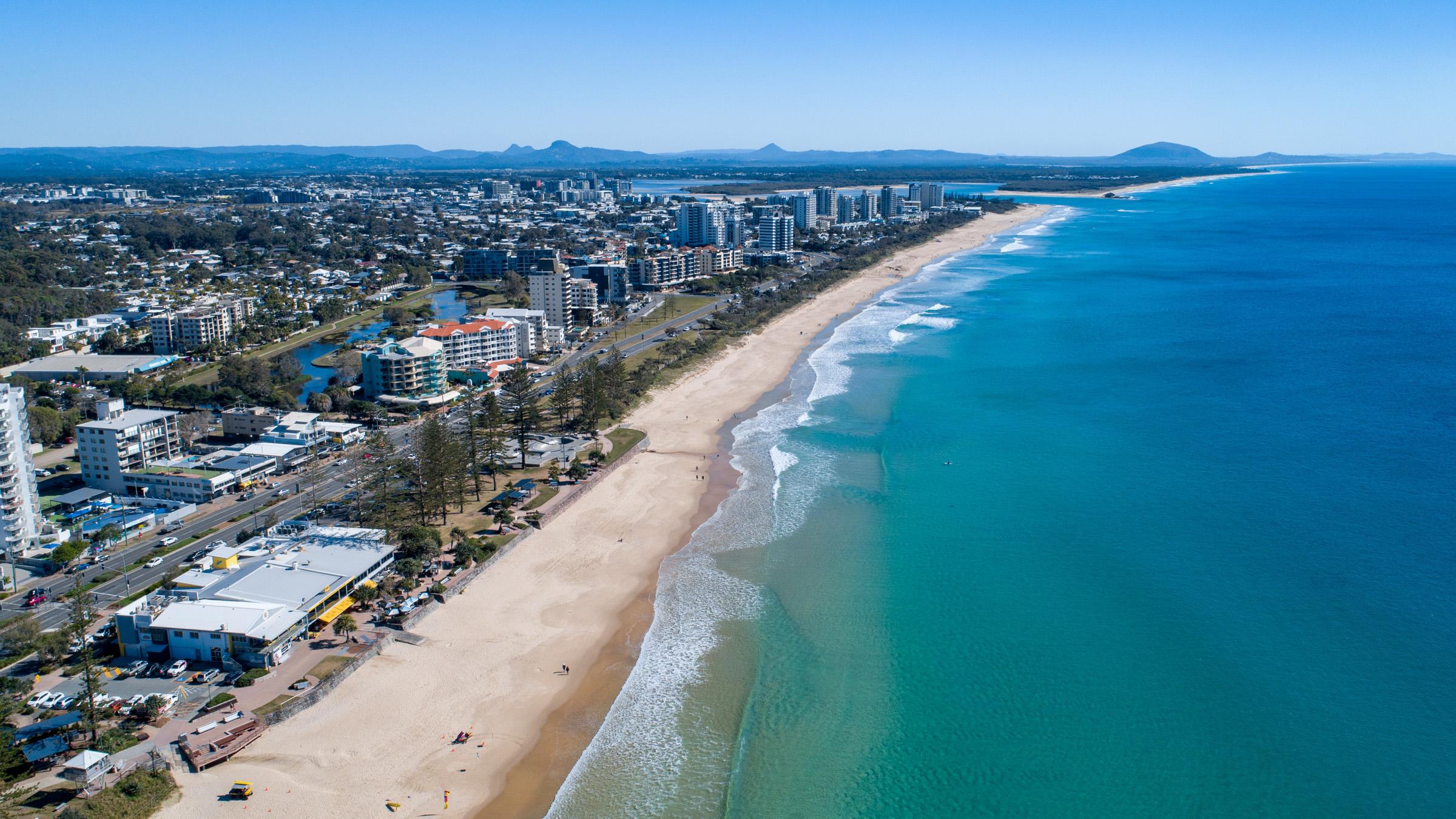 Aerial shot of Alexandra Coast Headland on a sunny day, with hotels, mountain ranges and surf lifesavers along the beach 