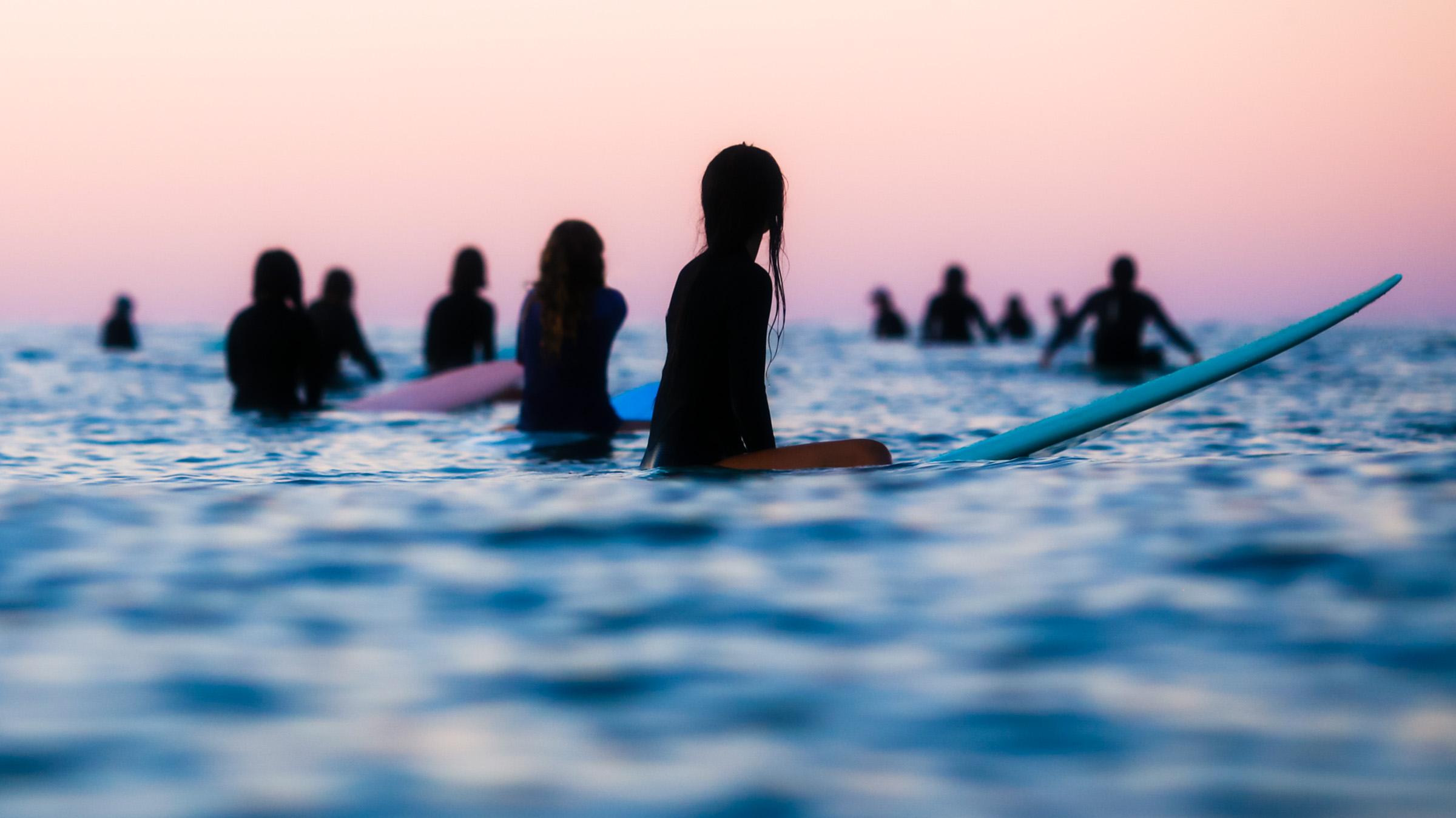 Group of surfers in the water silhouetted against a pink sky, waiting for a wave