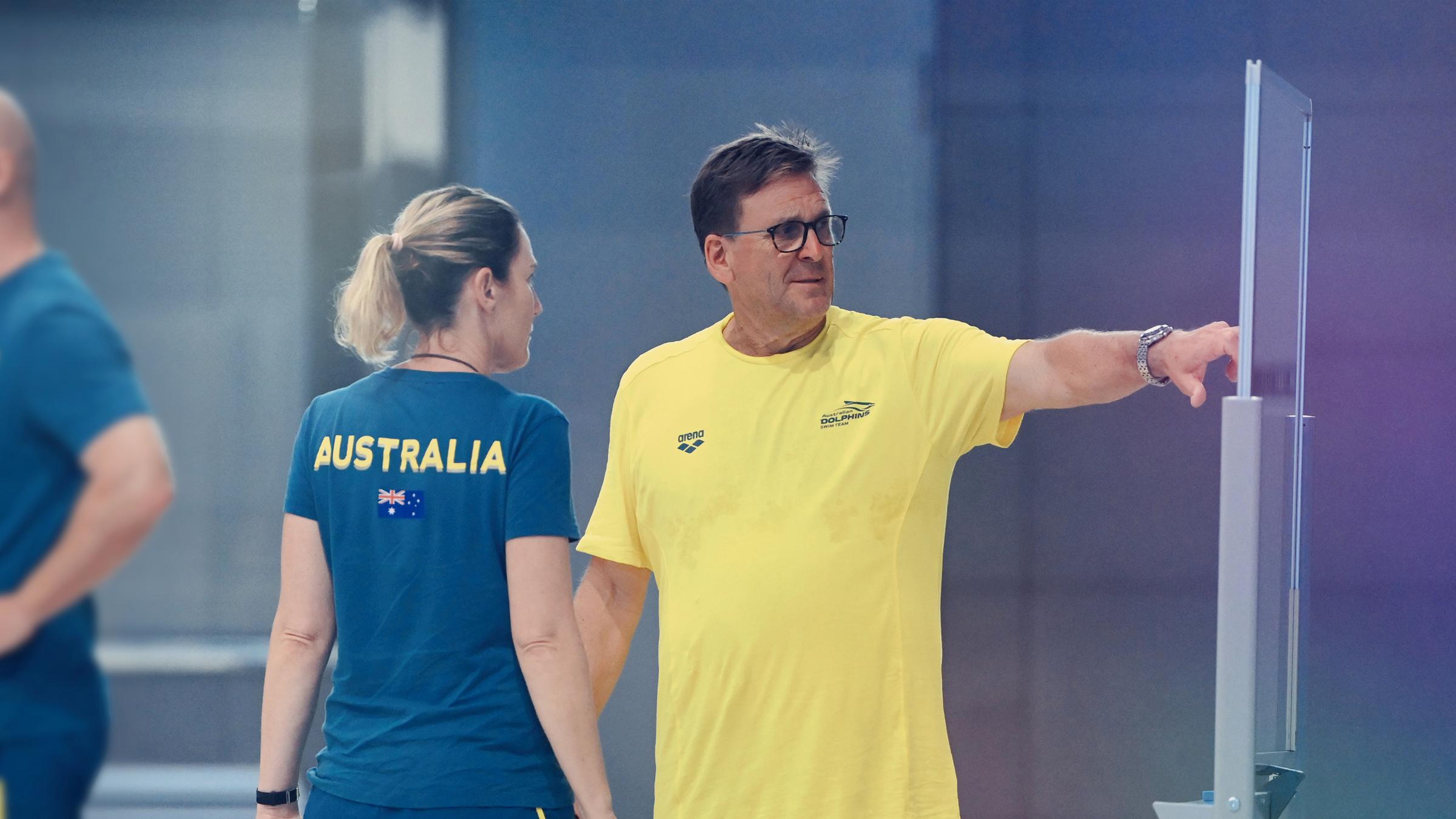 Michael Bohl is wearing a yellow t-shirt and green shorts. He is in the centre of the image pointing to a whiteboard on the right. Coach Amanda Issac (left) is listening to the planned session from Michael