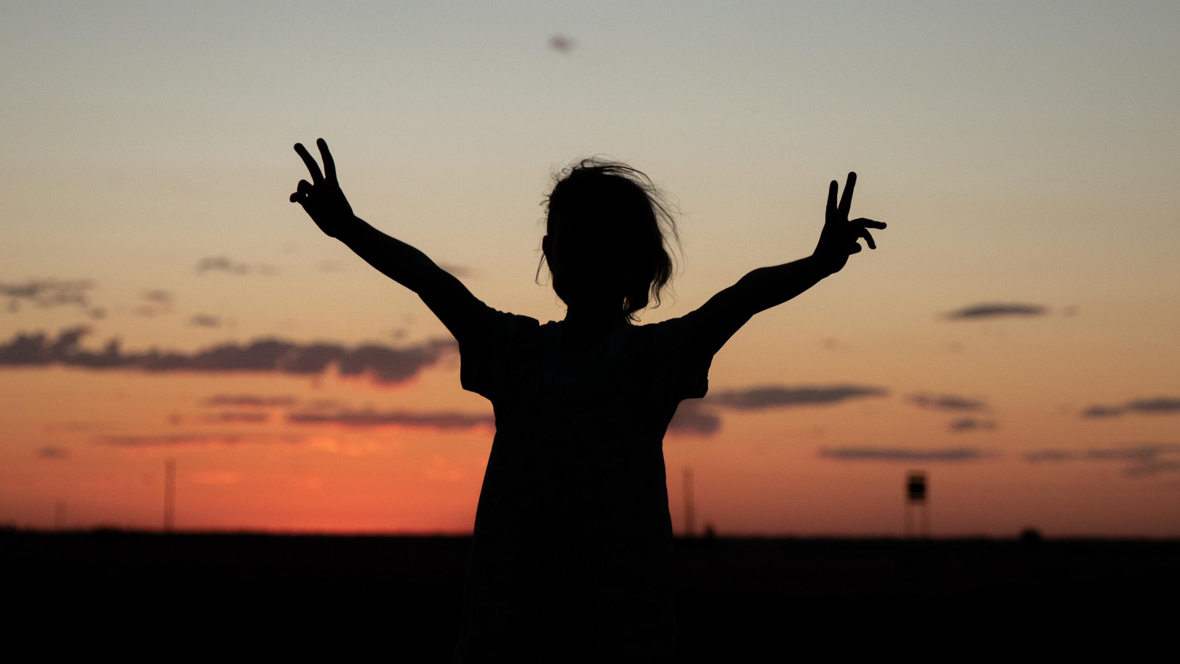 Child with outstretched arms silhouetted against a dark orange sunset