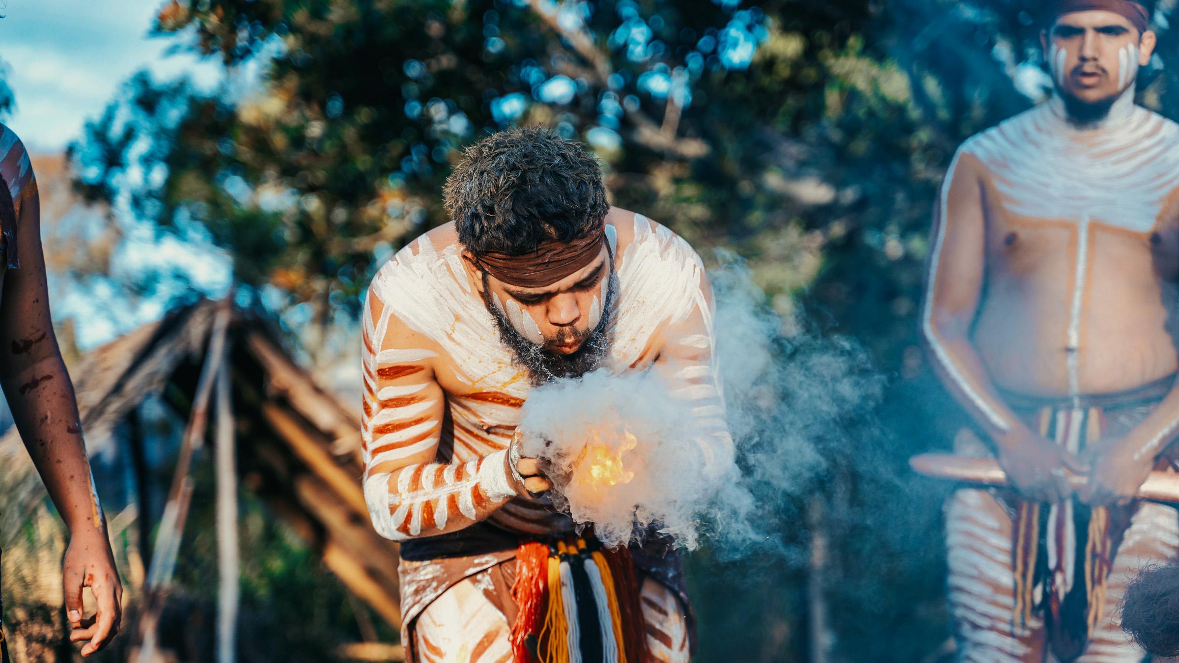 Young First Nations man with traditional face and body paint blowing onto a fire held in his hands