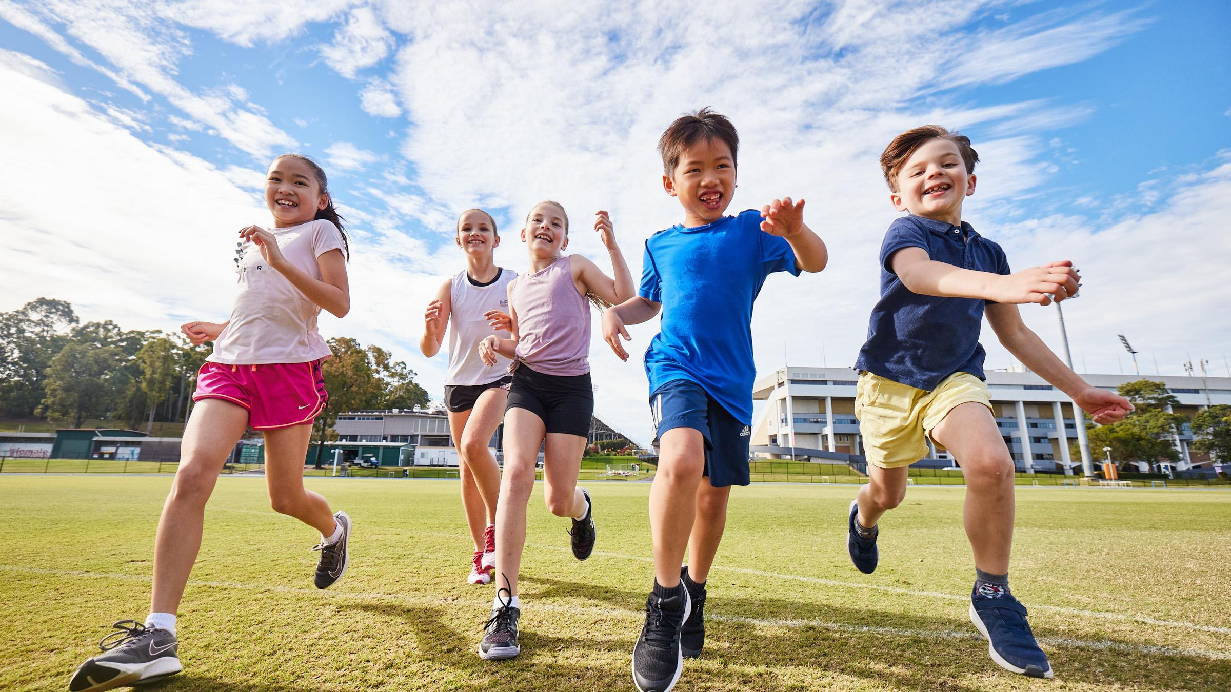 Five young children running across a green sports oval towards the camera