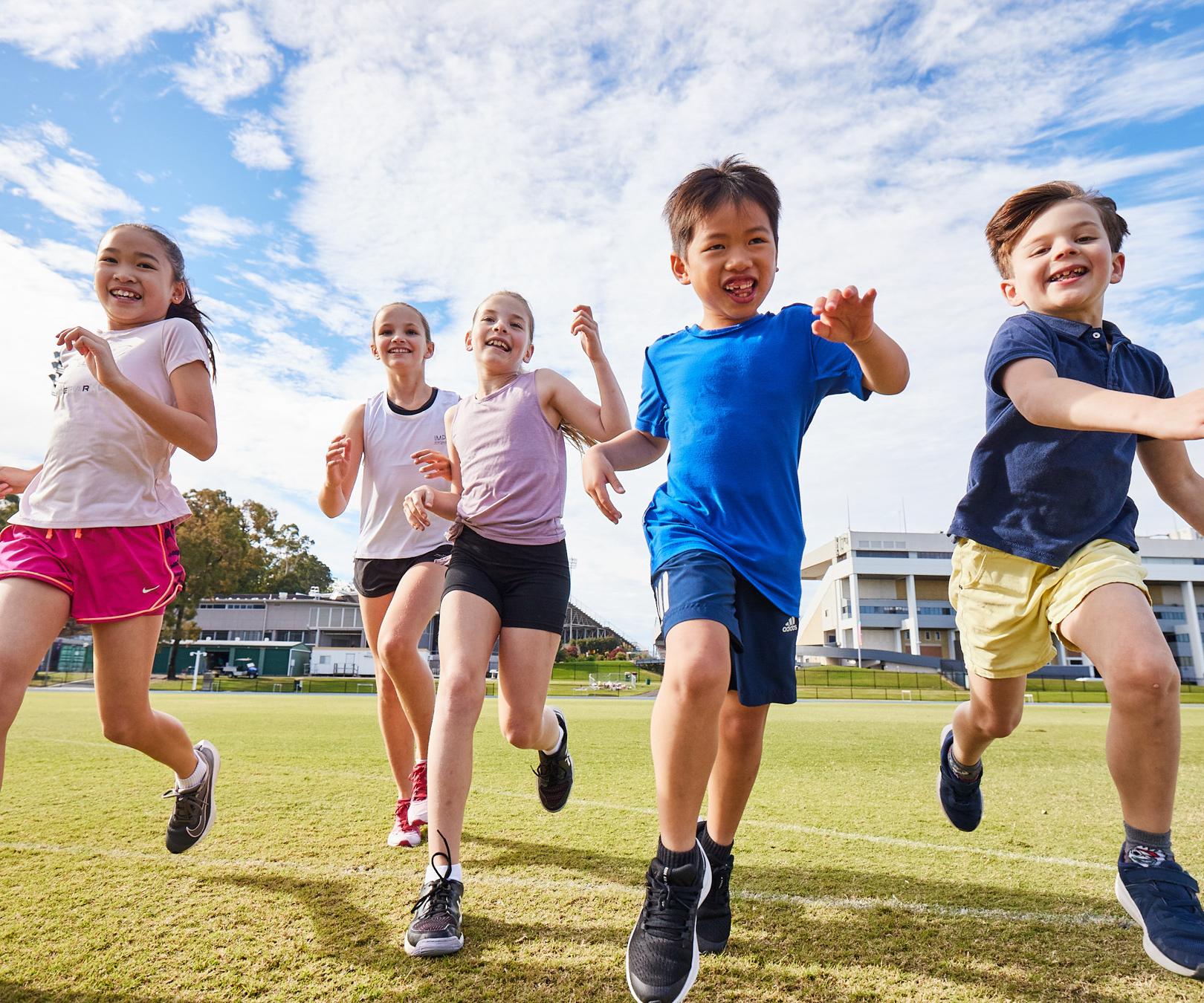 Five young children running across a green sports oval towards the camera