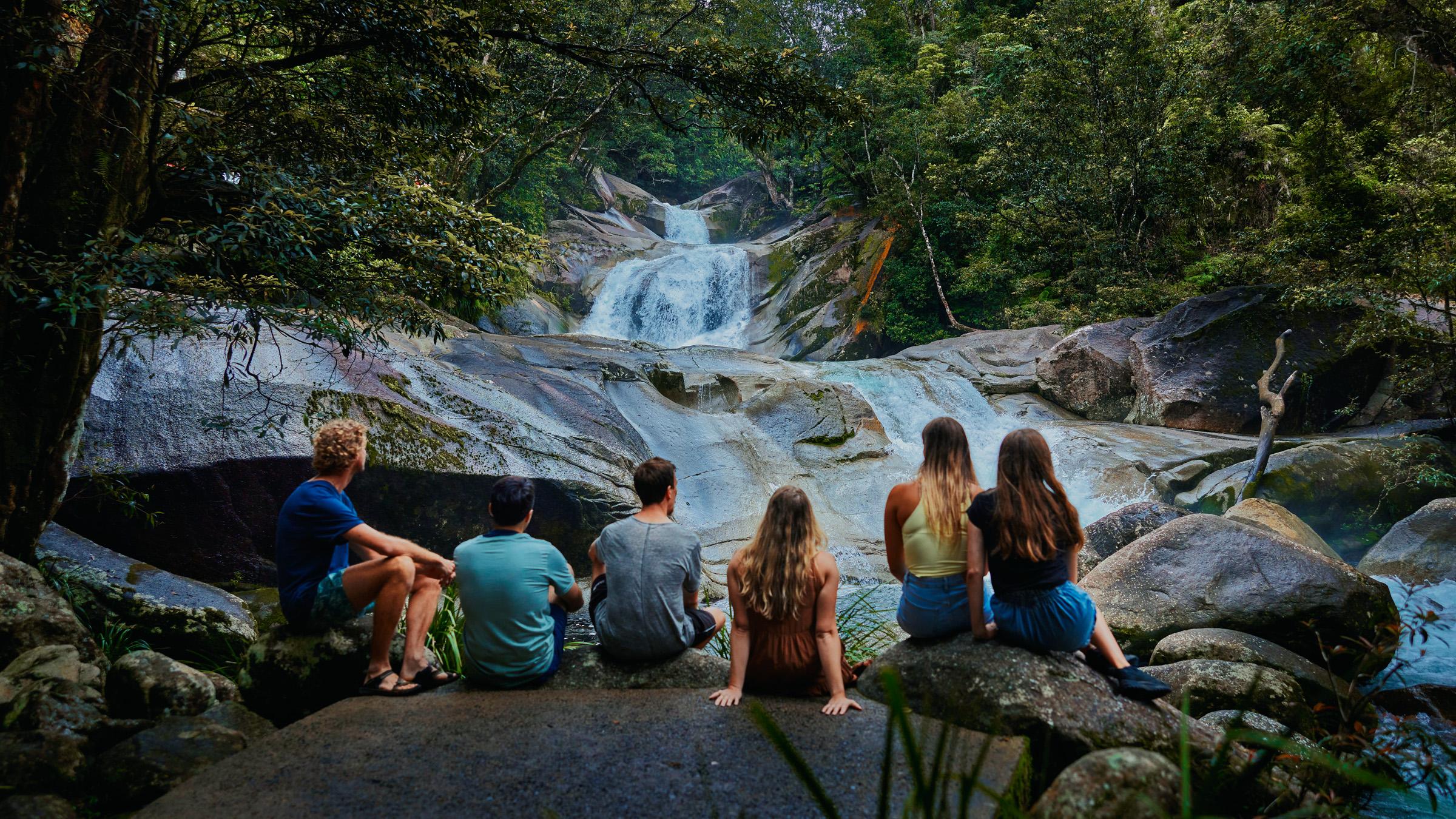 Group of people sitting on rocks at the bottom of a waterfall, surrounded by green, tropical rainforest