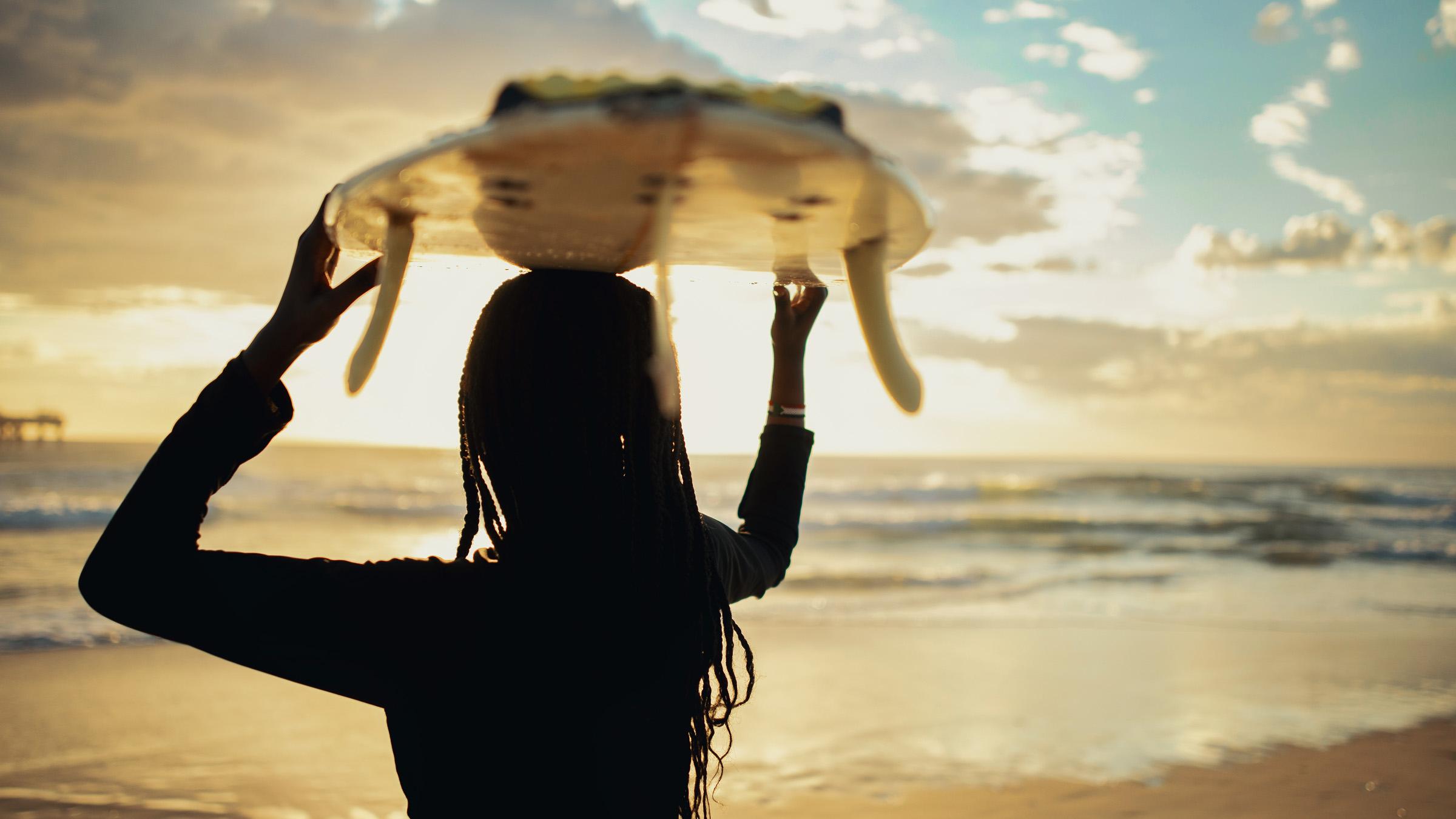 A young woman with braids and a surfboard looks out to the waves at the beach.