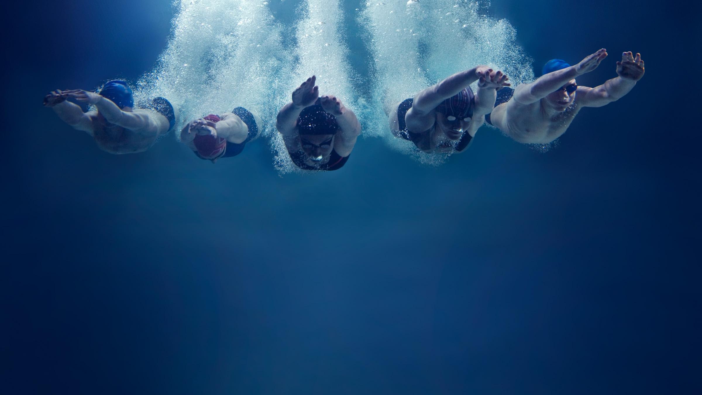 Underwater shot of five swimmers diving towards camera with a trail of bubbles behind them
